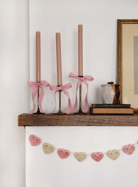 fireplace mantel with brass candlesticks on it with pink bows on candlesticks and heart garland hanging on mantel