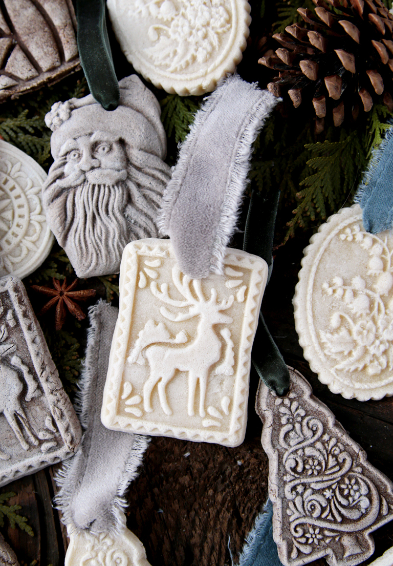 reindeer salt dough ornament laying next to christmas ornaments on wood board with ribbon and pinecone