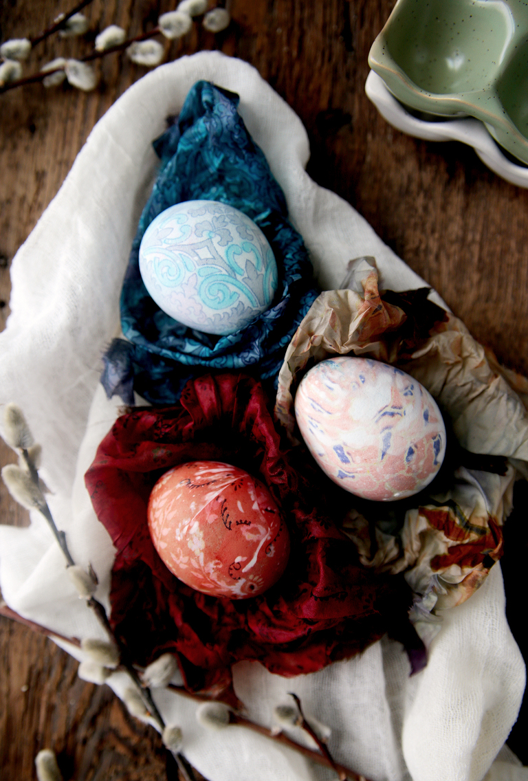 three dyed easter eggs sitting on silk fabric with pussy willows next to them