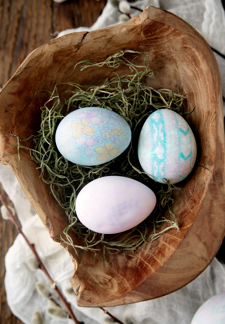 three dyed eggs in wood bowl with green moss sitting on white fabric with pussy willows next to it