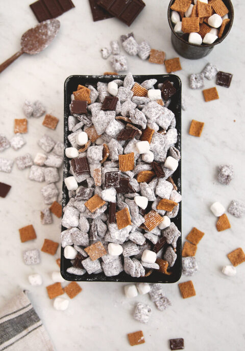 black loaf pan filled with s'more muddy buddies snack mix with cereal pieces and marshmallows scattered virtually pan