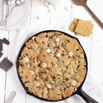 chocolate chip skillet cookie with marshmallows on white board with chocolate and graham crackers next to skillet