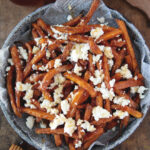 plate filled with sweet potato fries with goat cheese and hot honey on fries with honey stick and jar of honey next to plate
