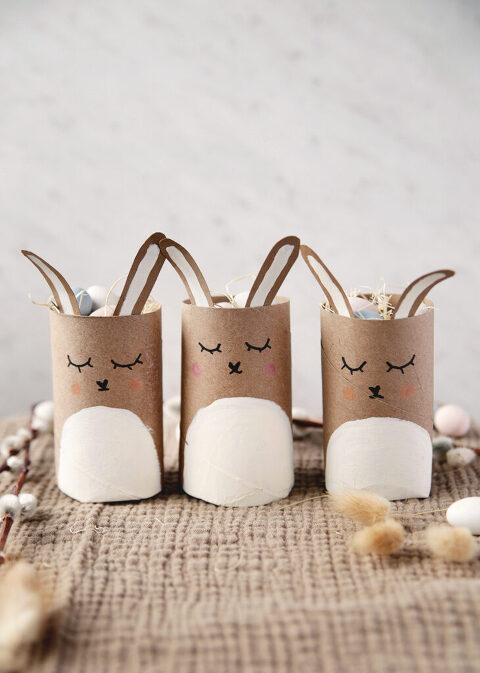 three toilet paper roll mini easter basket bunnies with candy eggs in them on brown cloth next to pussy willows and dried flowers