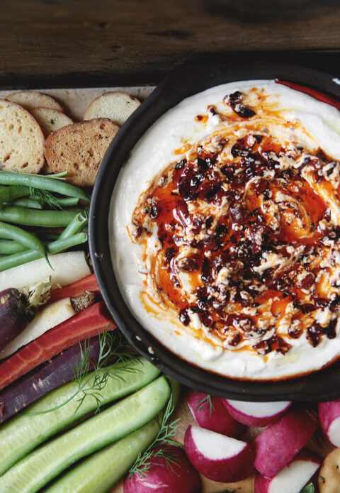 bowl of Whipped Cottage Cheese Chili Crisp Dip on cutting board with cut up vegetables and crackers surrounding it