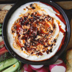black bowl with cottage cheese chili crisp dip in it surrounded by cut up vegetables on wood cutting board